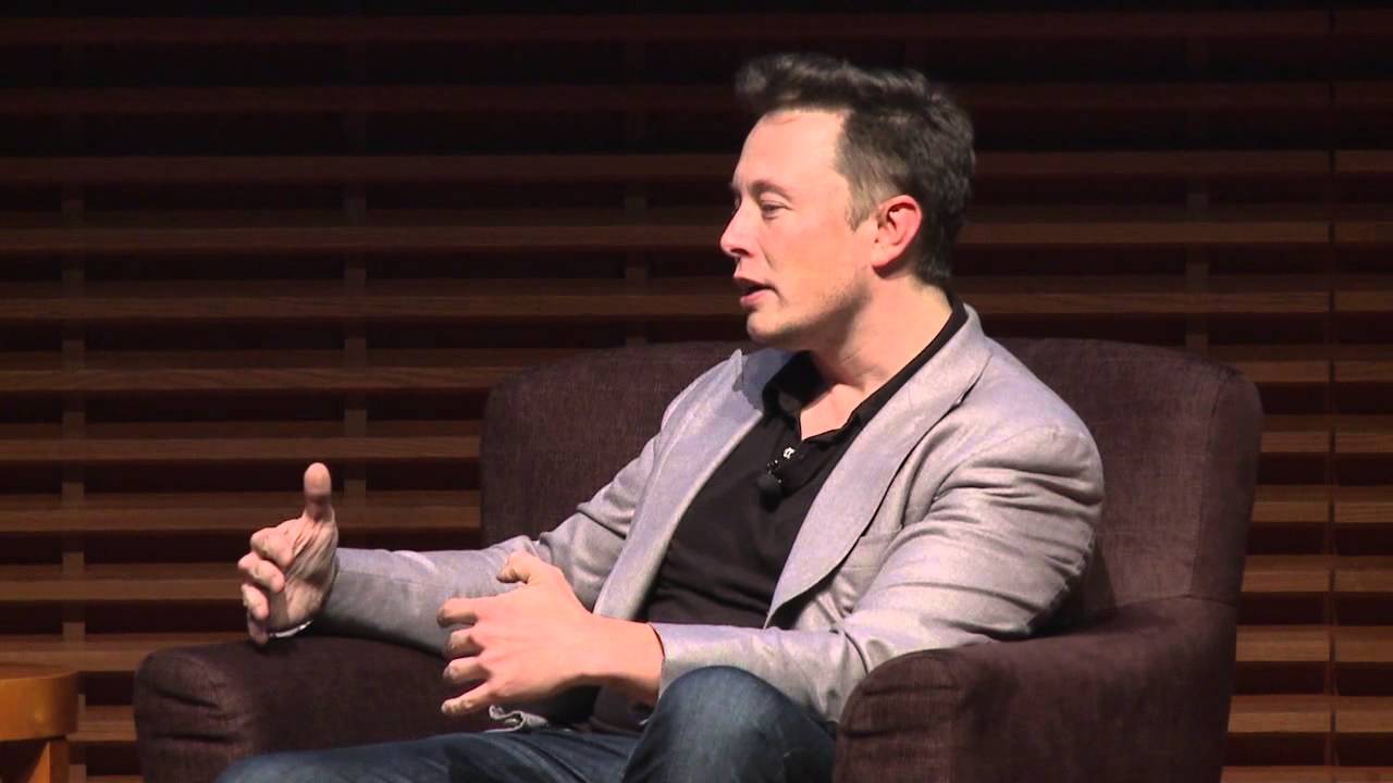 Elon Musk seated onstage in a light gray jacket. Official still from Stanford Graduate School of Business.